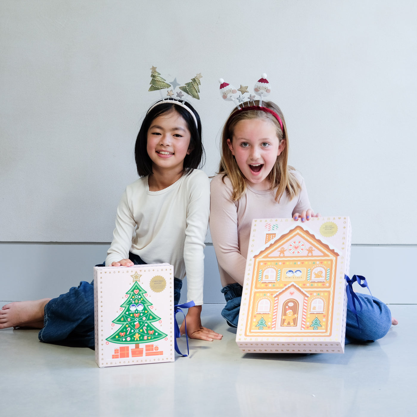 Two young girls wearing fun Christmas headbands exploring a medium and large sized cardboard advent calendar in pretty pink with blue satin ribbon tie, one with a gingerbread house design and the other with a green Christmas tree design, the calendar is filled with a joyful mix of children's accessories including hair accessories