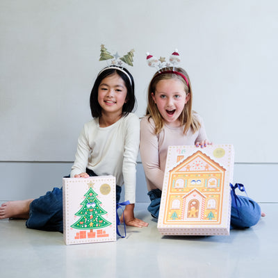 Two young girls wearing fun Christmas headbands exploring a medium and large sized cardboard advent calendar in pretty pink with blue satin ribbon tie, one with a gingerbread house design and the other with a green Christmas tree design, the calendar is filled with a joyful mix of children's accessories including hair accessories
