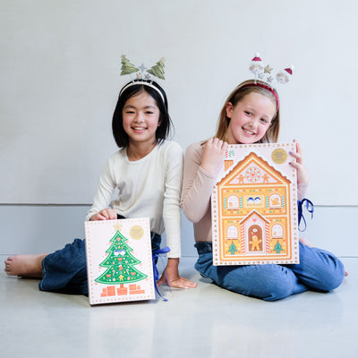 Two young girls wearing fun Christmas headbands exploring a medium and large sized cardboard advent calendar in pretty pink with blue satin ribbon tie, one with a gingerbread house design and the other with a green Christmas tree design, the calendar is filled with a joyful mix of children's accessories including hair accessories