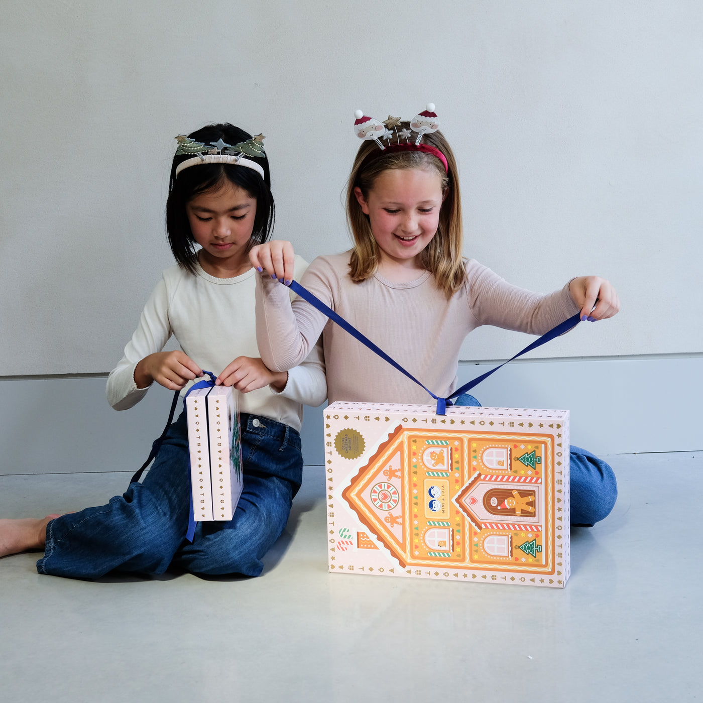 Two young girls wearing fun Christmas headbands exploring a medium and large sized cardboard advent calendar in pretty pink with blue satin ribbon tie, the calendar is filled with a joyful mix of children's accessories including hair accessories