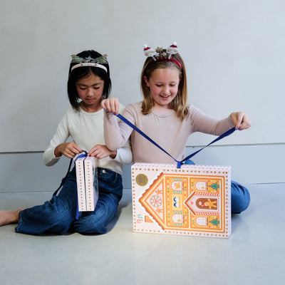Two young girls wearing fun Christmas headbands exploring a medium and large sized cardboard advent calendar in pretty pink with blue satin ribbon tie, the calendar is filled with a joyful mix of children's accessories including hair accessories