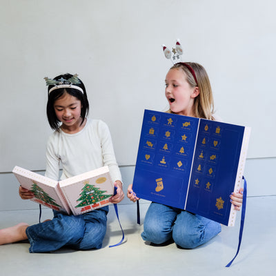 Two young girls wearing fun Christmas headbands exploring a medium and large sized cardboard advent calendar in pretty pink with blue satin ribbon tie, one with a gingerbread house design and the other with a green Christmas tree design, the calendar is filled with a joyful mix of children's accessories including hair accessories