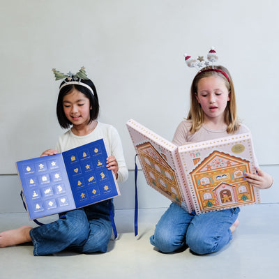 Two young girls wearing fun Christmas headbands exploring a medium and large sized cardboard advent calendar in pretty pink with blue satin ribbon tie, one with a gingerbread house design and the other with a green Christmas tree design, the calendar is filled with a joyful mix of children's accessories including hair accessories