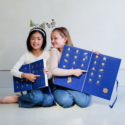 Two young girls wearing fun Christmas headbands exploring a medium and large sized cardboard advent calendar in pretty pink with blue satin ribbon tie, one with a gingerbread house design and the other with a green Christmas tree design, the calendar is filled with a joyful mix of children's accessories including hair accessories