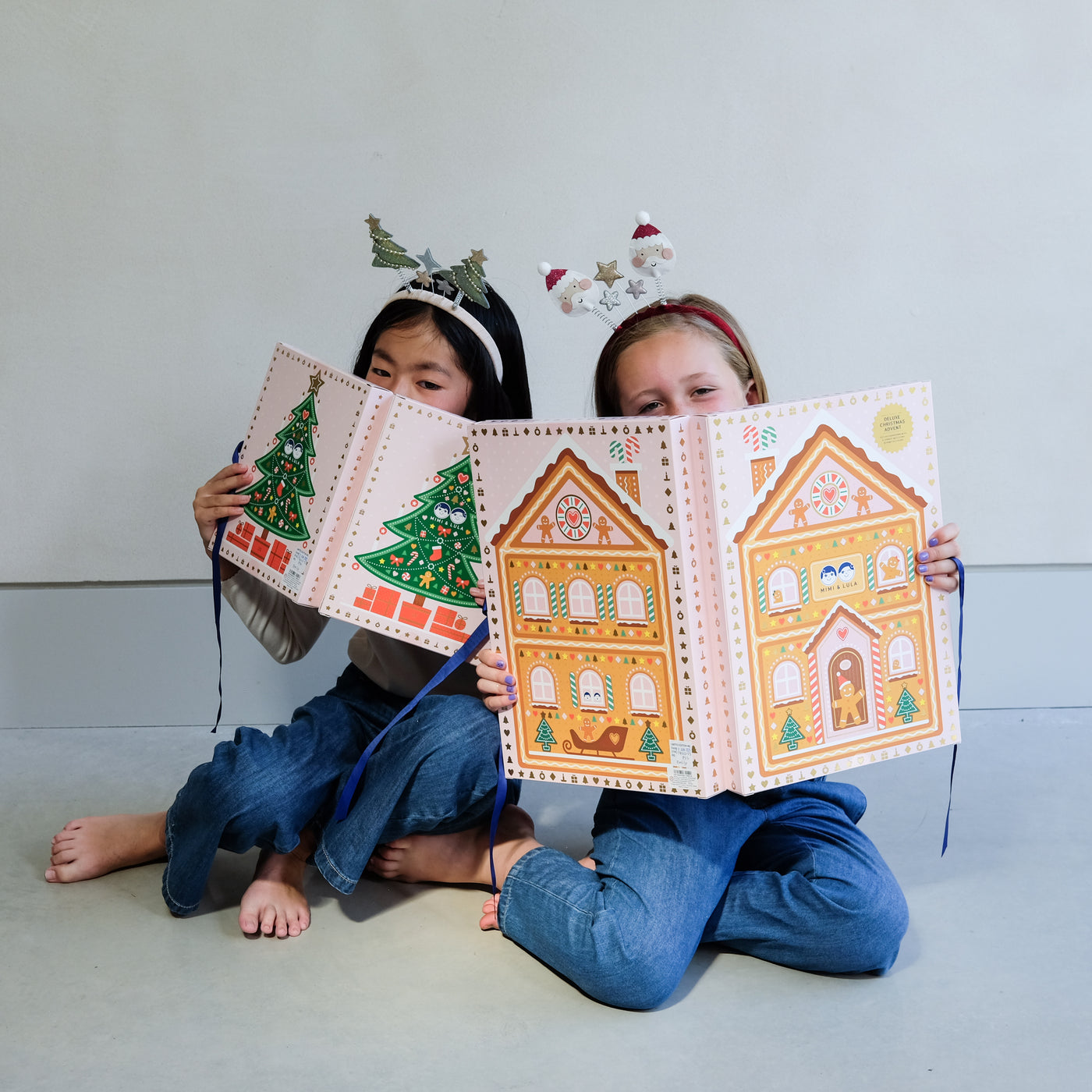 Two young girls wearing fun Christmas headbands exploring a medium and large sized cardboard advent calendar in pretty pink with blue satin ribbon tie, one with a gingerbread house design and the other with a green Christmas tree design, the calendar is filled with a joyful mix of children's accessories including hair accessories
