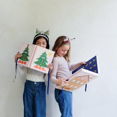 Two young girls wearing fun Christmas headbands exploring a medium and large sized cardboard advent calendar in pretty pink with blue satin ribbon tie, one with a gingerbread house design and the other with a green Christmas tree design, the calendar is filled with a joyful mix of children's accessories including hair accessories