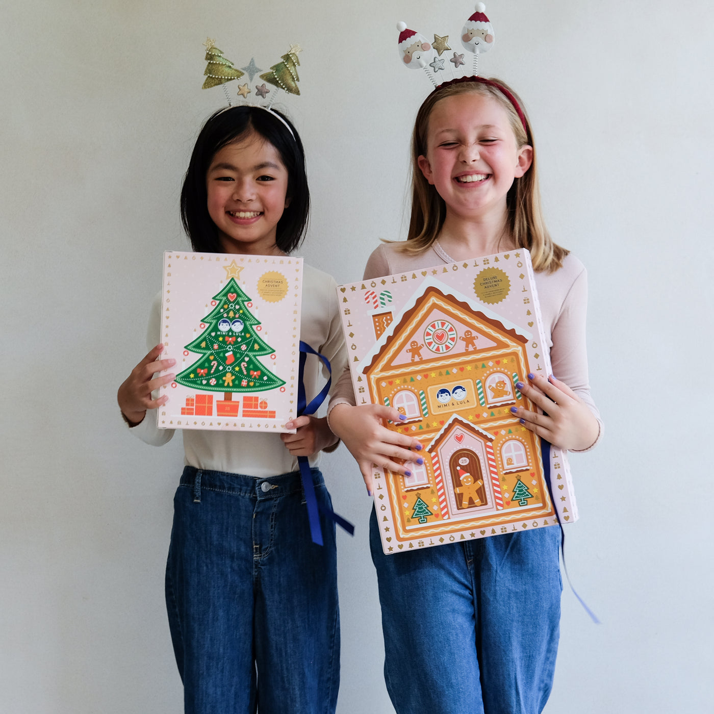 Two young girls wearing fun Christmas headbands exploring a medium and large sized cardboard advent calendar in pretty pink with blue satin ribbon tie, one with a gingerbread house design and the other with a green Christmas tree design, the calendar is filled with a joyful mix of children's accessories including hair accessories